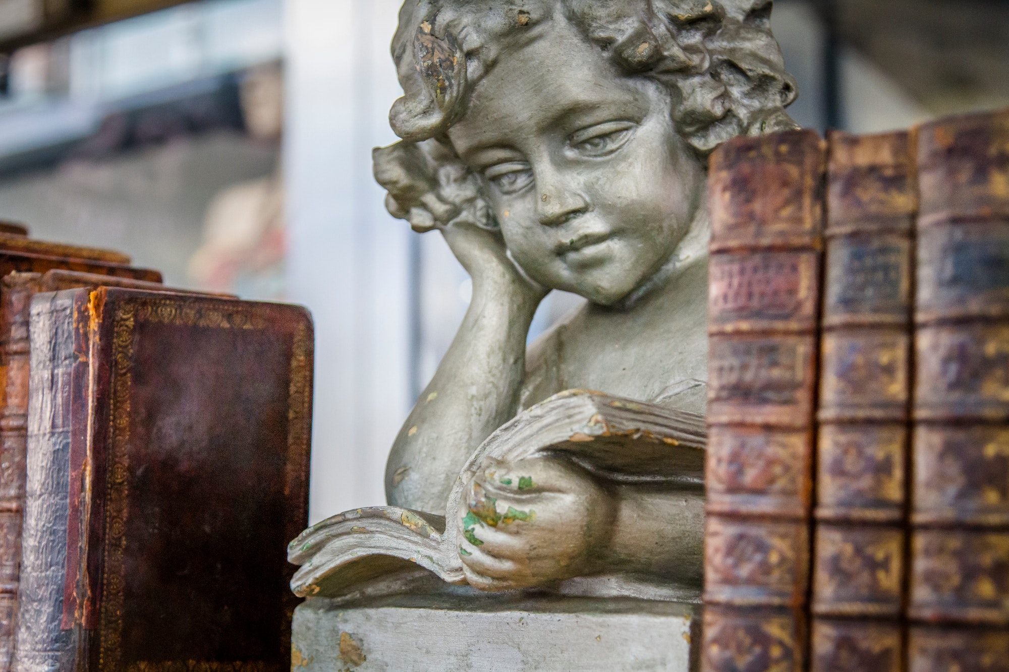 Angelic boy angel statue reading a book between a stack of old books, outside market for sale.