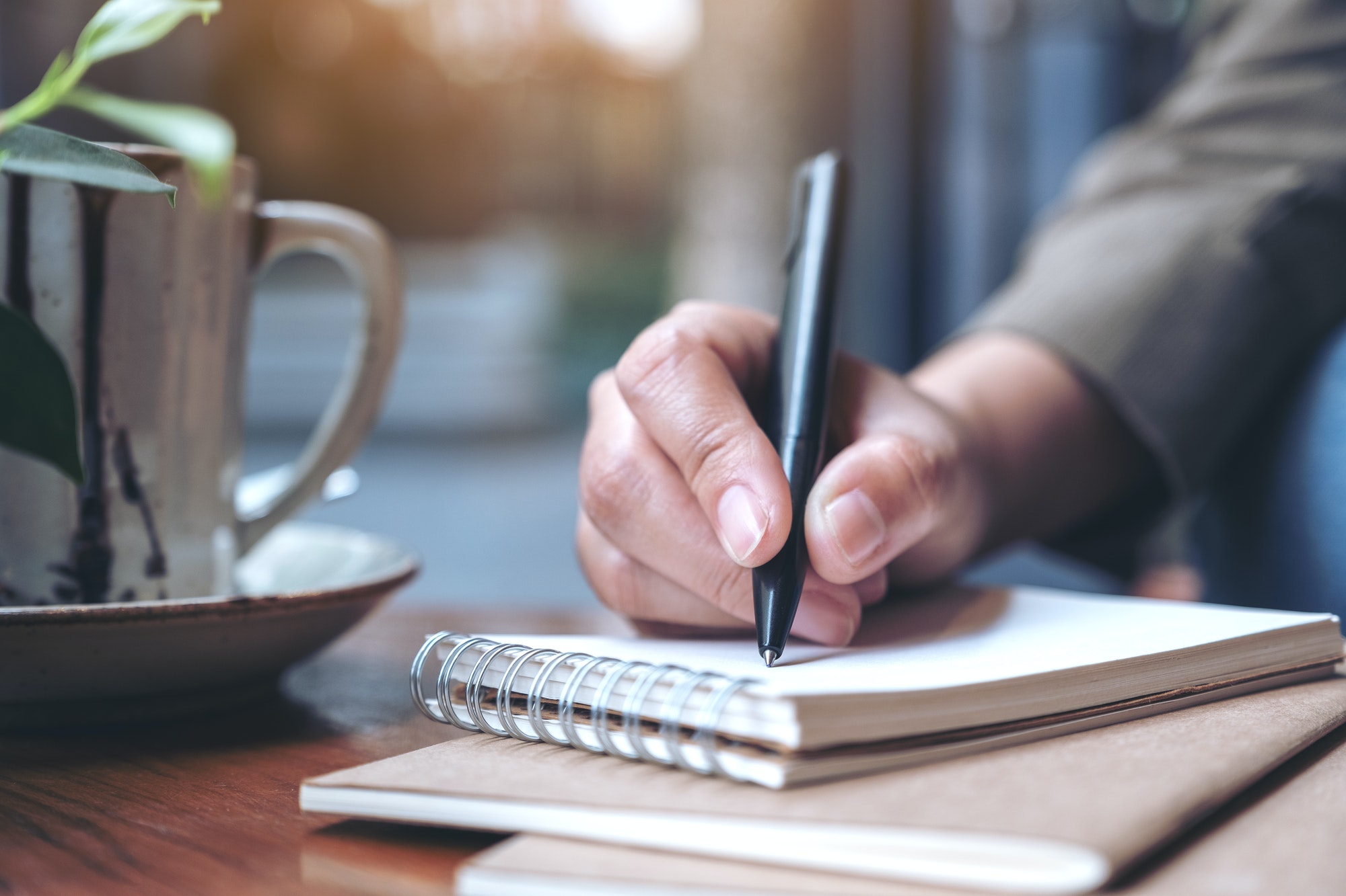 Closeup image of a woman's hand writing on blank notebook with coffee cup on wooden table